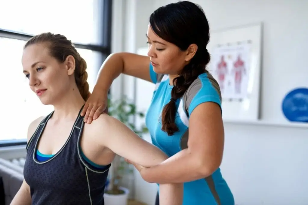 Mulher com o cabelo loiro trançado usando top de academia preto, sendo avaliada por médica com o cabelo preto trançado usando camiseta azul