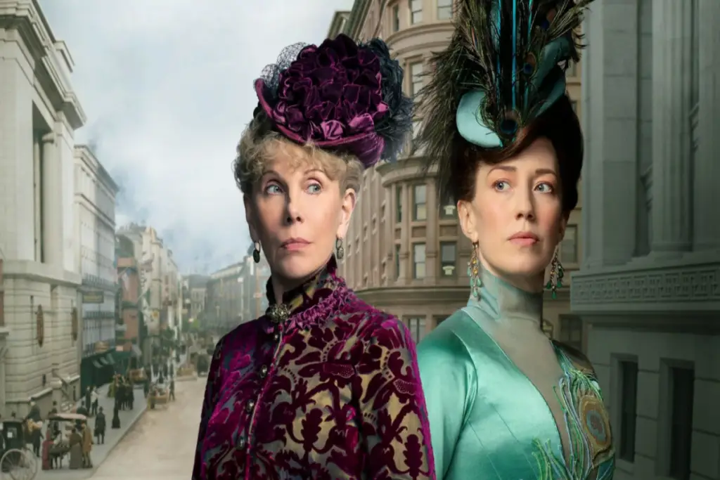 Duas mulheres uma ao lado da outra e atrás uma imagem da cidade de Nova York, ambas estão vestidas com roupas de época, uma em tons de roxo e outra em tons de verde