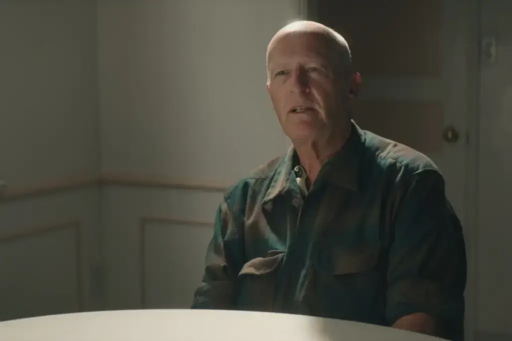 Homem careca usando uma roupa marrom e verde sentado em frente a uma mesa