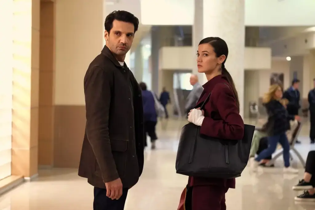 Homem com cabelo curto, usando blazer marrom e calça e blusa preta parado ao lado de uma mulher com o cabelo amarrado em rabo de cavalo e usando uma roupa social vinho com um curativo na mão e com uma bolsa preta