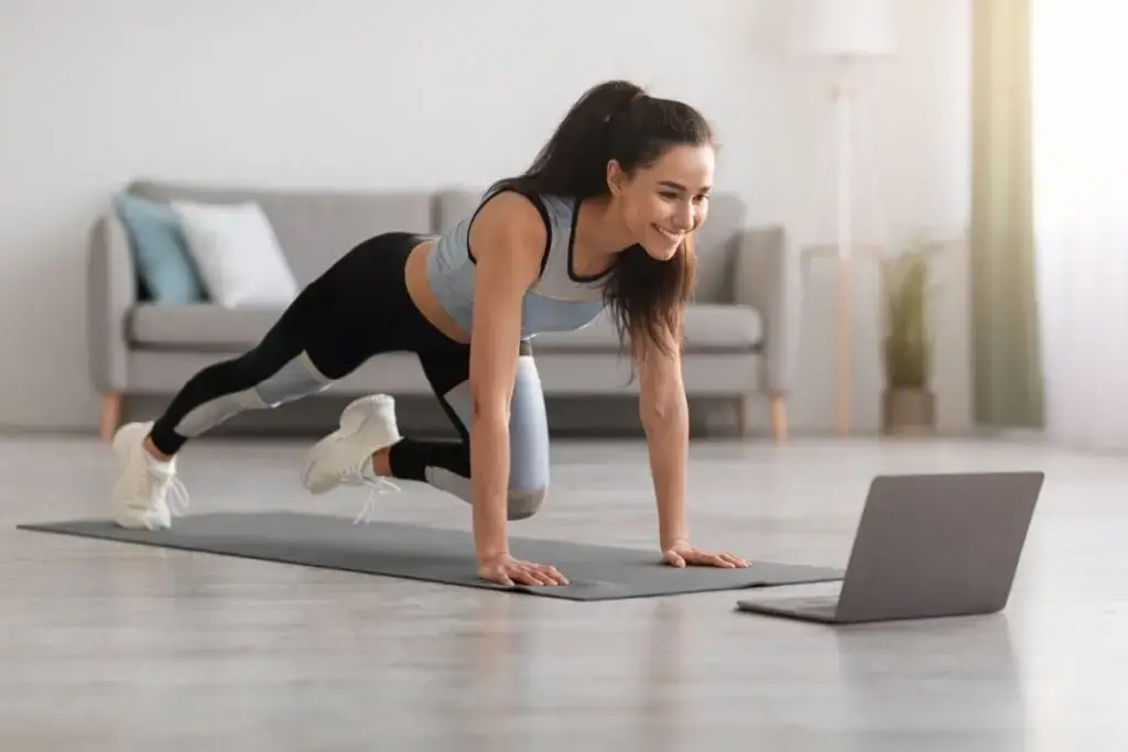 Mulher com roupa de academia praticando exercício na sala de casa, assistindo a um vídeo no notebook