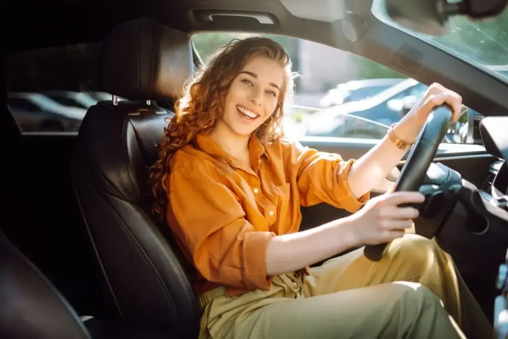 mulher de blusa laranja e calça amarela sentada dentro de carro e sorrindo