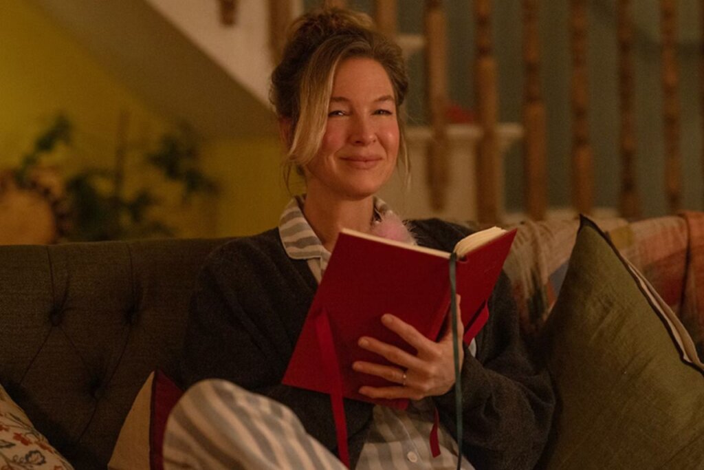 Cena do filme "Bridget Jones: Louca pelo Garoto" com Bridget sentada em um sofá, sorrindo e segurando um livro