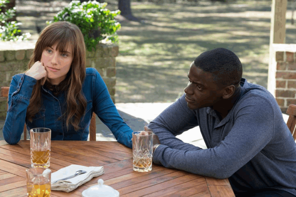 Homem negro e mulher branca sentados em uma mesa de madeira ao ar livre, com copos de bebida à frente