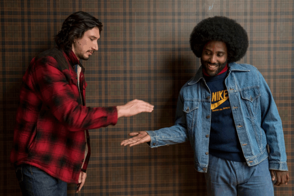 Dois homens, um de barba e casaco vermelho e outro de cabelo black e jaqueta jeans, fazendo um cumprimento amigável