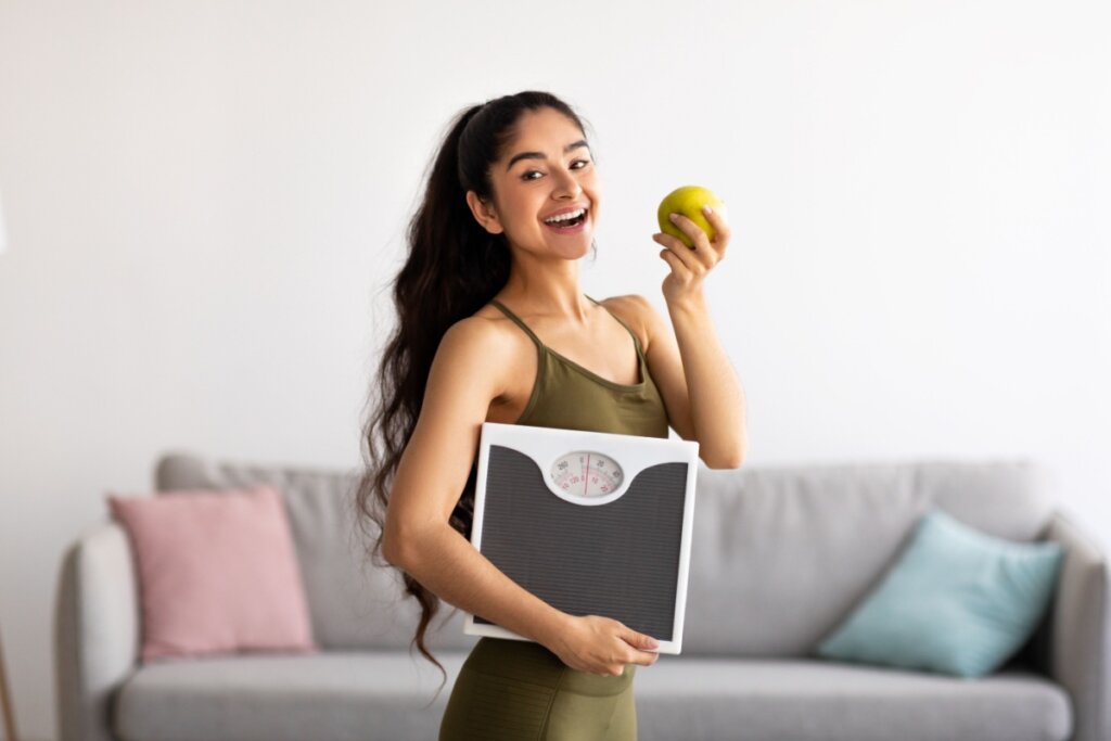 Jovem com cabelo comprido encaracolado preso em rabo de cavalo, usando roupa de academia verde, segurando uma balança em uma mão e uma maçã-verde na outra