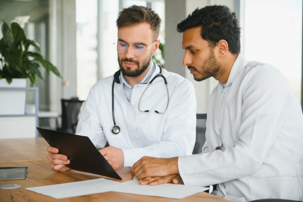 médico e paciente vestidos de branco, sentados lado a lado e olhando para prancheta preta apoiada em mesa