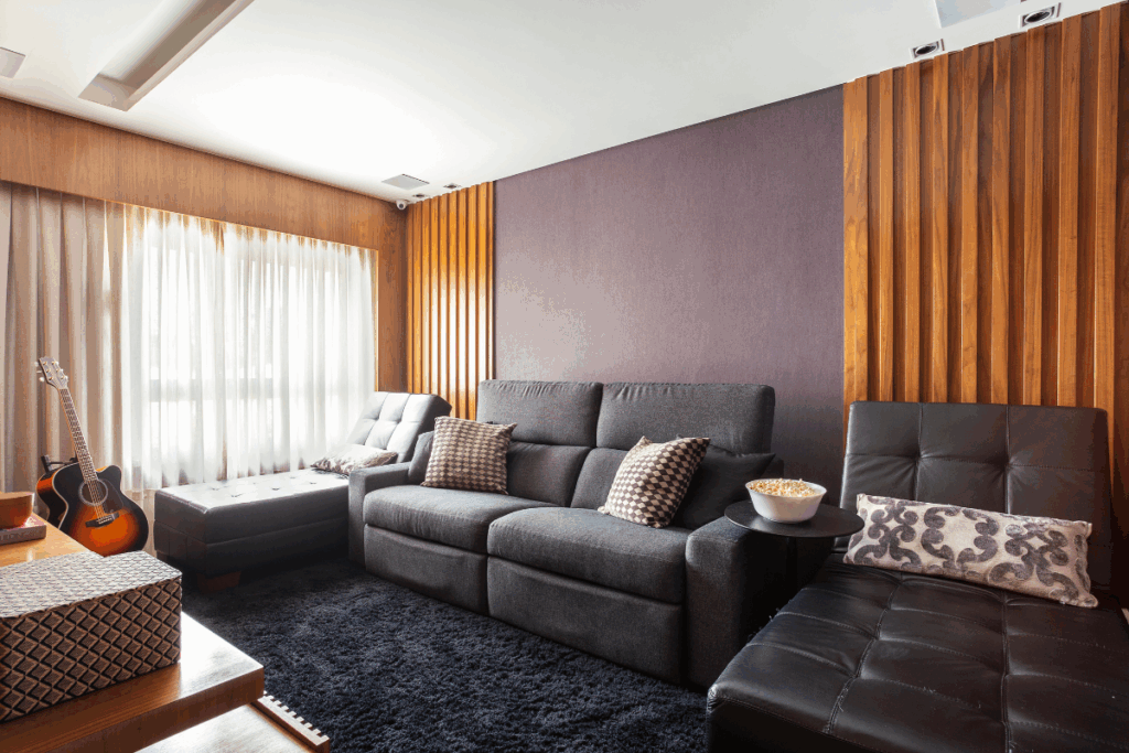 Sala de estar com tapete preto, parede cinza e em madeira, com sofá cinza com almofadas bege e poltrona preta