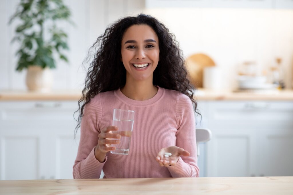 mulher de cabelo cacheado e blusa rosa sorrindo e segurando copo de água e comprimidos