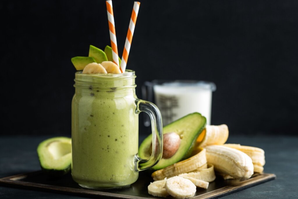 Vitamina de abacate com banana servida em copo de vidro, com metade de abacate ao lado e uma banana e rodelas de banana em cima de bancada preta em fundo preto