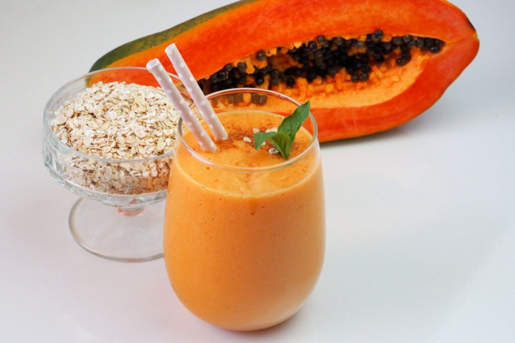 Vitamina de mamão com aveia servido em copo de vidro com metade de um mamão ao lado e uma tigela com aveia