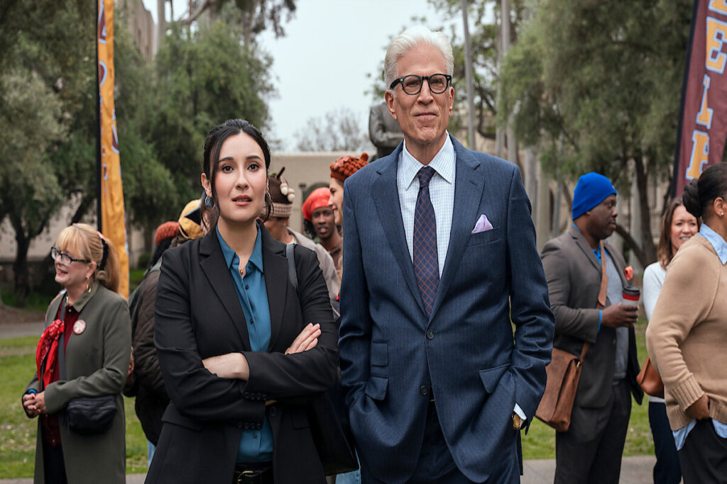 mulher baixa de terno ao lado de homem alto de terno e cabelo braco com pessoas ao fundo