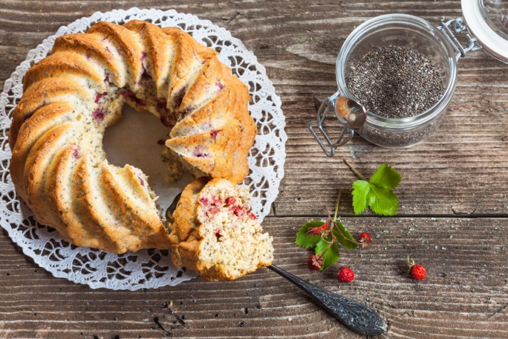 Bolo de chia com morangos com um pedaço cortado ao lado de recipiente com chia e morangos em superfície amadeirada