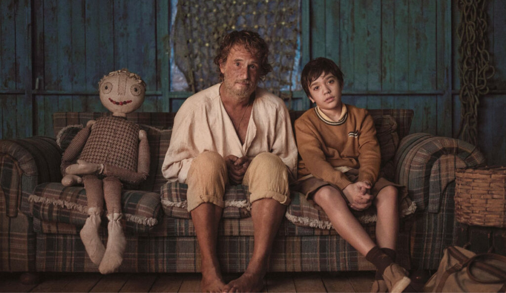 homem de camiseta branca sentado em sofá ao lado de menino de blusa marrom e boneco