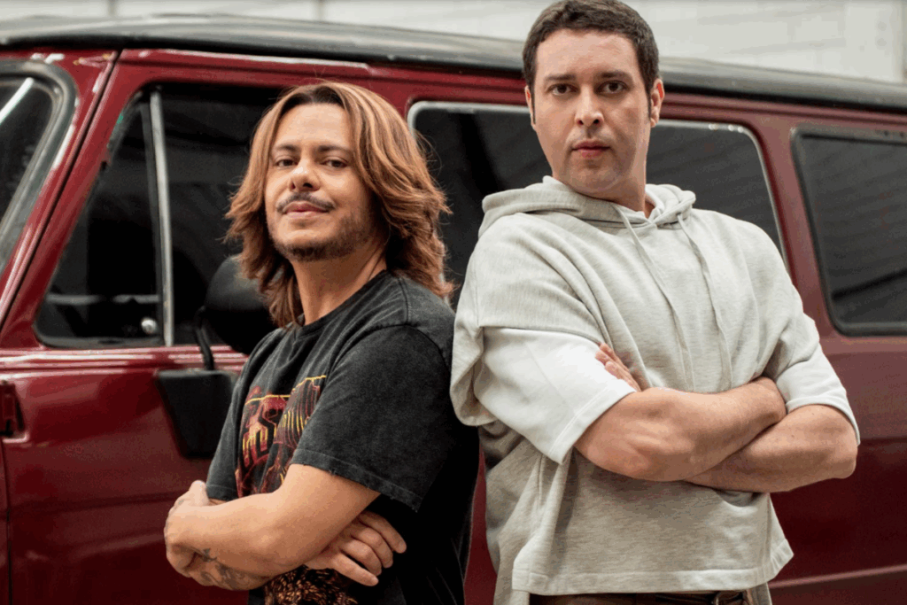 Dois homens posando lado a lado com braços cruzados em frente a um veículo vermelho