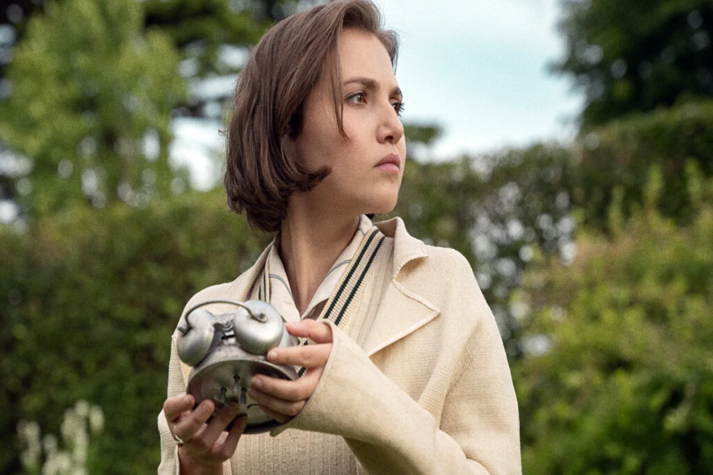 Jovem segurando um objeto metálico com relógios enquanto observa algo fora de cena em um ambiente arborizado