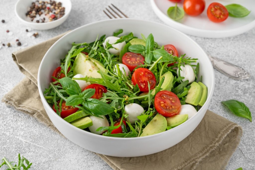 Recipiente branco com folhas de rúcula, pedaços de abacate, tomate e queijo de búfala 