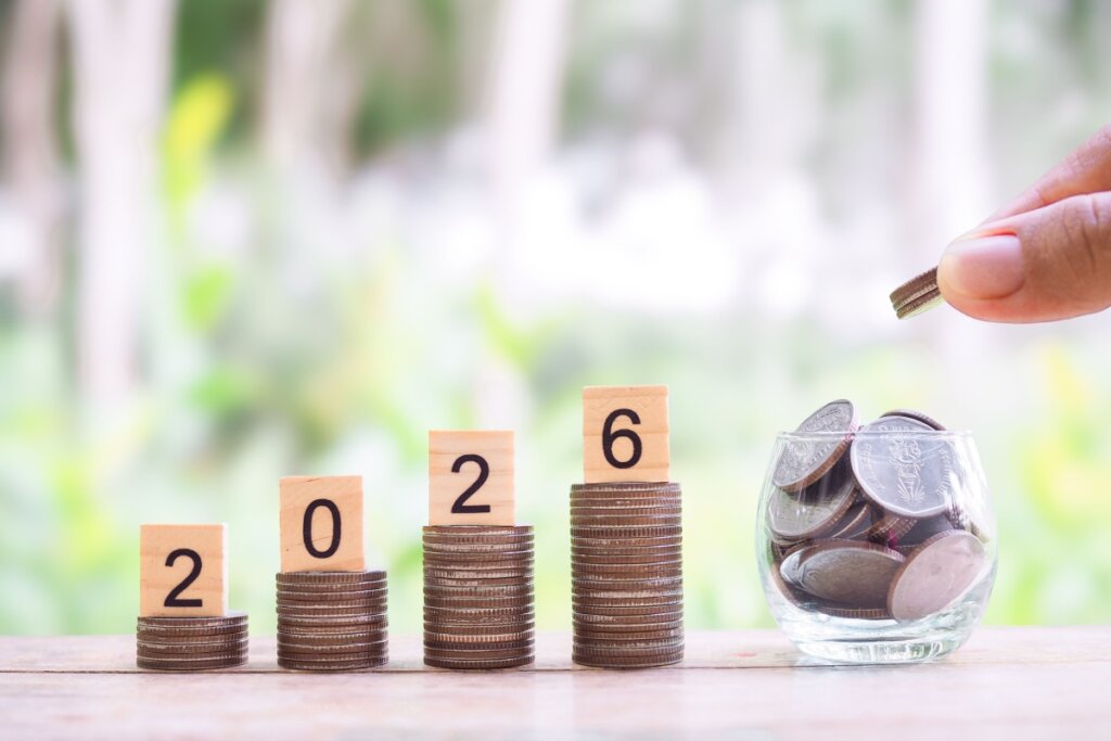 Blocos de madeira com os números 2026 em cima de pilhas de moedas. Pote transparente com moedas ao lado 