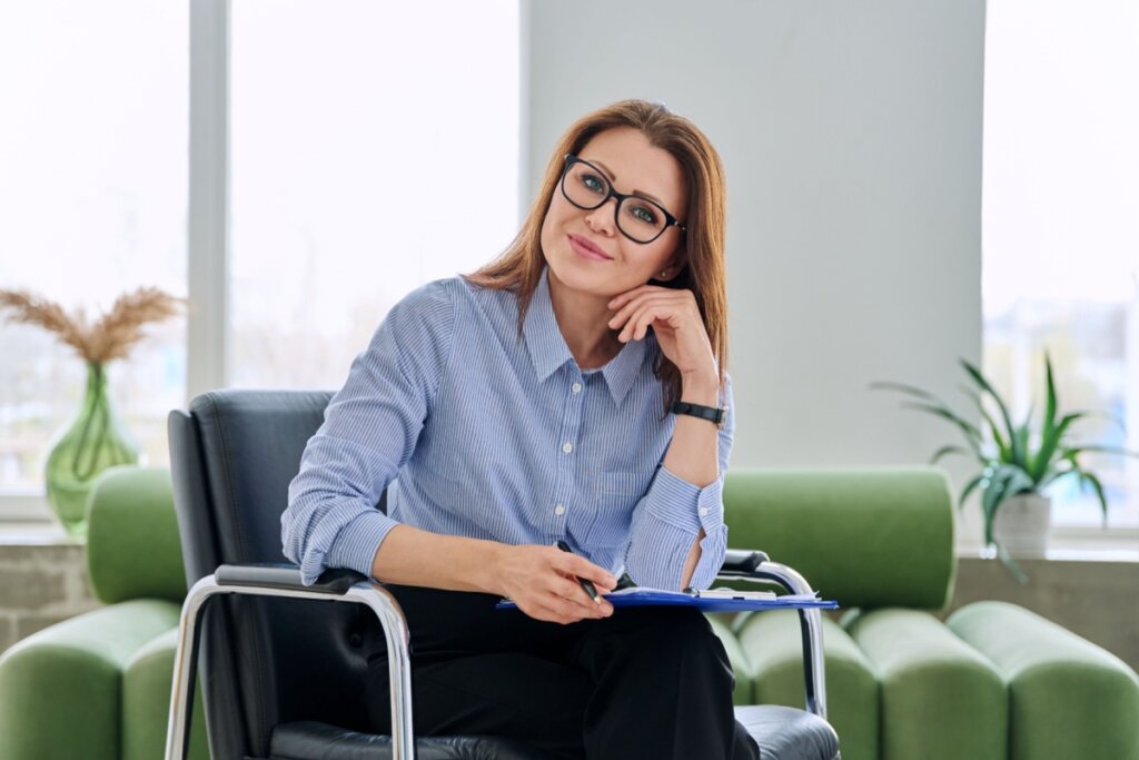 Mulher com o cabelo loiro e solto, usando camisa de botões azul sentada em cadaeira em consultório