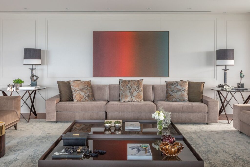 Sala de estar com quadro colorido em uma parede branca e sofá bege-escuro ao fundo. Na frente do sofá, mesa de centro preta com objetos 