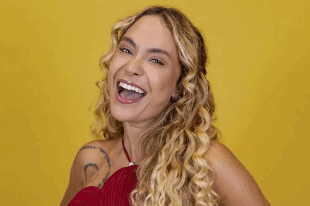 Mulher loira de cabelos ondulados sorrindo em fundo mostarda